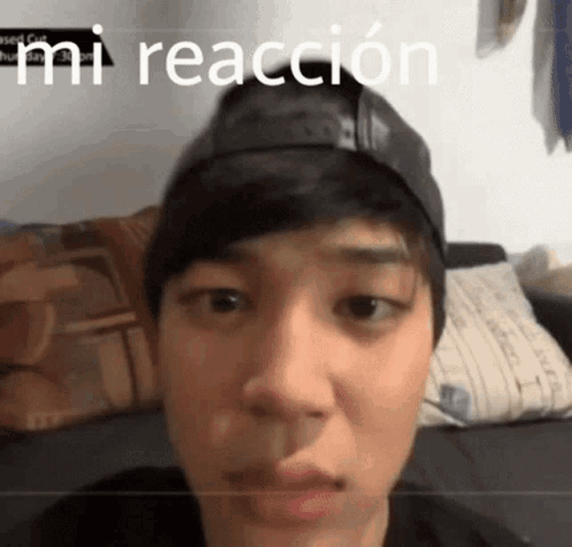 a man wearing a baseball cap is sitting on a couch with the words " mi reaccion " above his head .