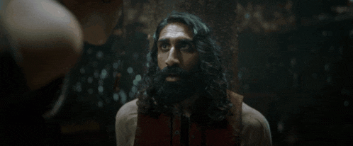 a man with long hair and a beard is talking to another man in a dark room