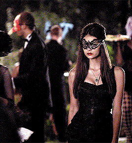 a woman wearing a black mask and a black dress is standing in a crowd of people .