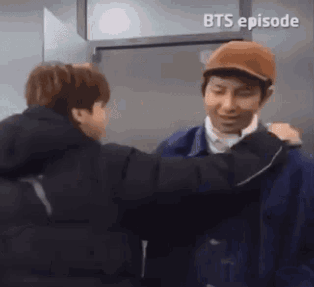 two men are hugging each other in front of a door . one of the men is wearing a hat .