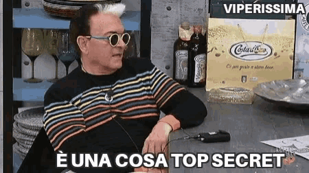 a man wearing sunglasses and a striped sweater is sitting at a table with the words e una cosa top secret below him