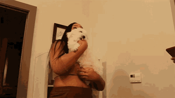 a woman holds a small white dog in her arms in front of a thermostat