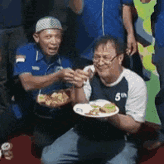 a man wearing a shirt that says 5th is feeding another man a plate of food