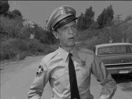 a black and white photo of a police officer in a uniform and tie standing on a road next to a car .