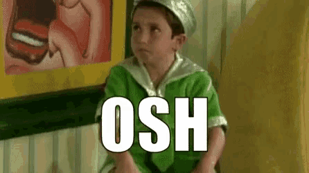 a young boy in a green sailor costume is sitting in a chair with the word osh written on the wall behind him