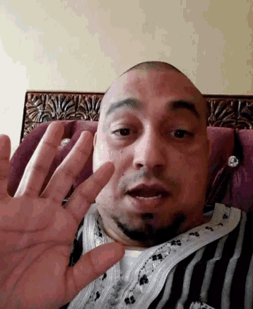 a bald man with a beard is sitting on a couch with his hand up .