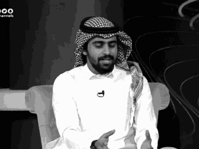 a man wearing a head scarf and a white shirt is sitting in front of a tv screen that says channels