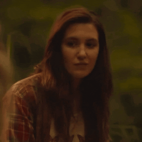 a woman with long red hair is wearing a plaid shirt and looking at the camera .