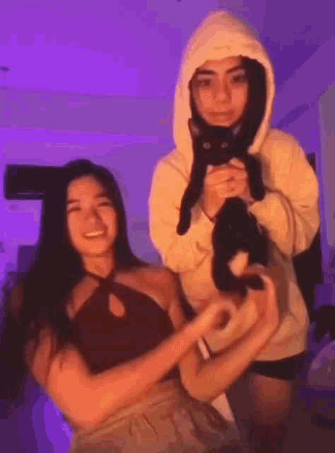 two girls are holding a black cat in their arms and making a heart shape with their hands .