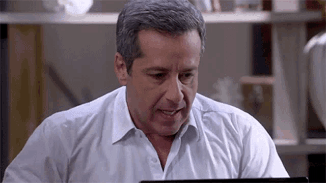 a man in a white shirt is sitting at a table looking at a laptop computer .