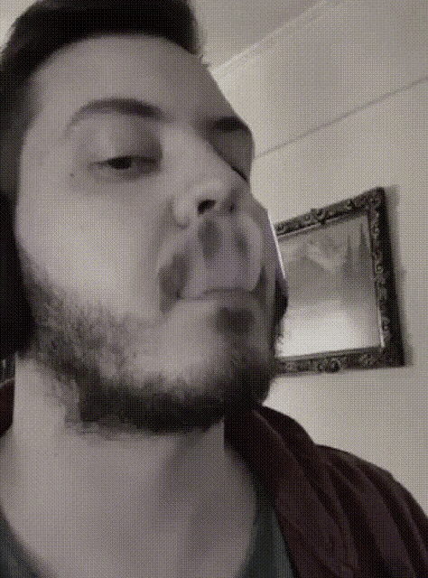 a man with a beard is smoking a cigarette in front of a framed picture on the wall