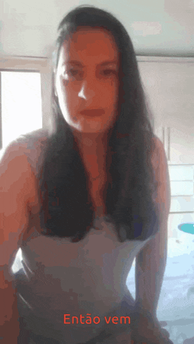 a woman in a white tank top is standing in front of a window with the words então vem written on the bottom