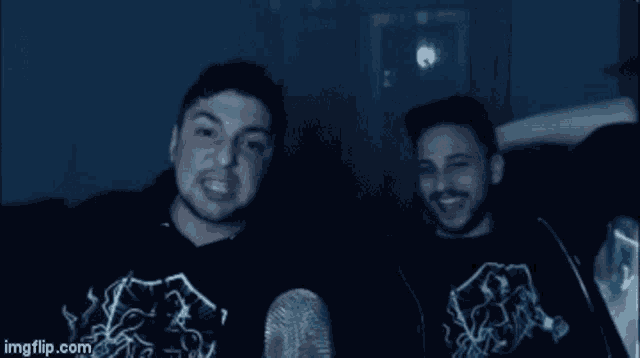 two men are sitting next to each other in a dark room wearing sweatshirts .