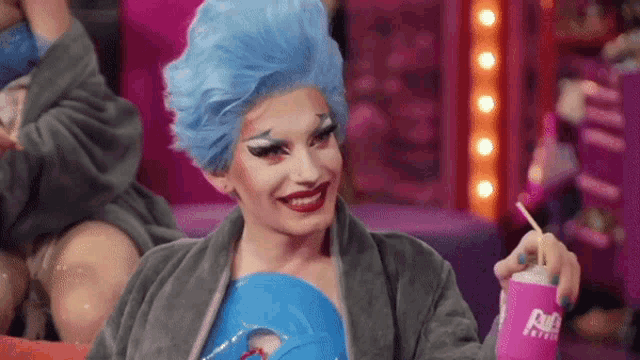 a drag queen with blue hair is smiling while holding a blue mask and a can of soda .