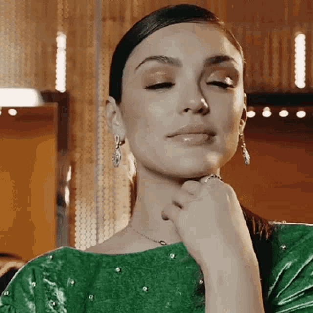 a woman wearing a green dress and earrings is looking down with her hand on her chin .