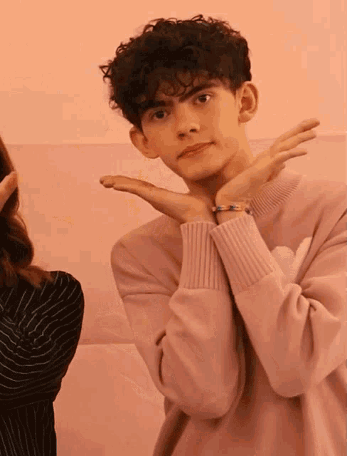 a man with curly hair wearing a pink sweater is making a heart shape with his hands