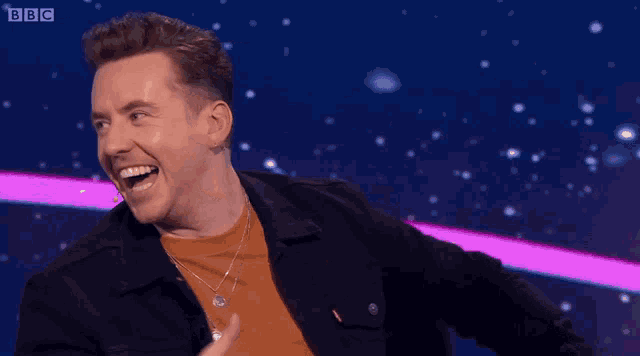 a man is laughing on a stage with bbc written on the bottom