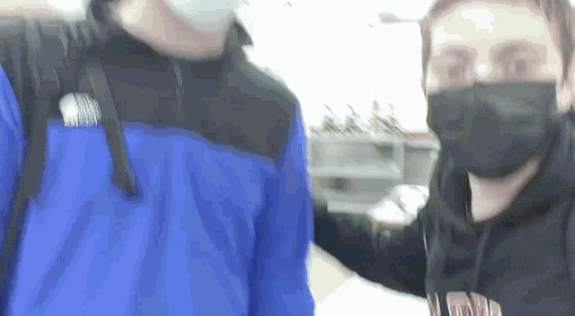 two men wearing face masks are standing next to each other . one of the men is wearing a blue jacket .
