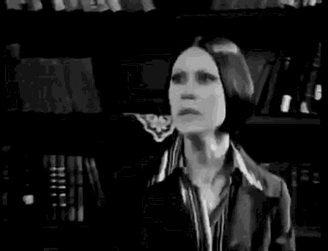 a black and white photo of a woman in a suit standing in front of a bookshelf .