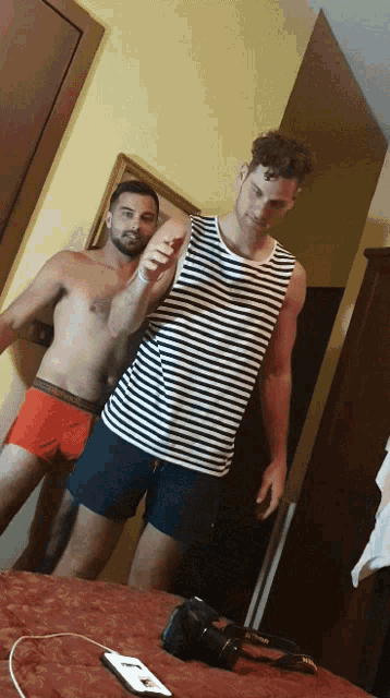 a man in a striped tank top stands next to another man in red boxer shorts