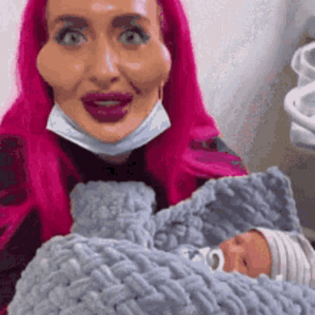a woman with pink hair is holding a baby wrapped in a blanket .