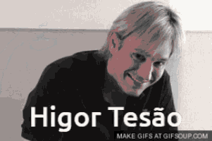 a man in a black shirt is smiling in front of a wall with the words higor tesão written on it .
