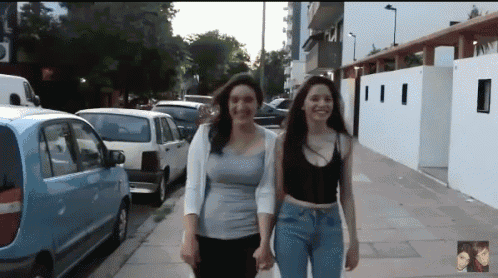 two women walking down a street holding hands with a blue car in the background