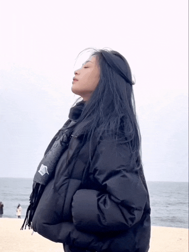 a woman wearing a black jacket and scarf stands on a beach