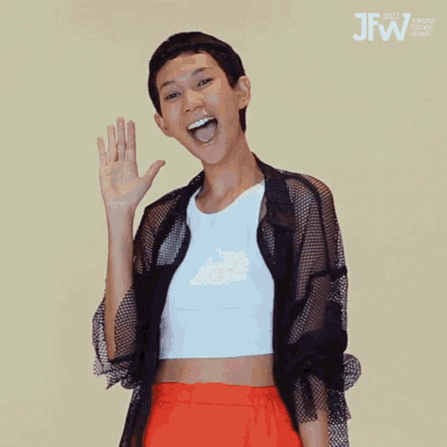 a woman wearing a white crop top and red shorts waves her hand in front of a sign that says jakarta fashion week