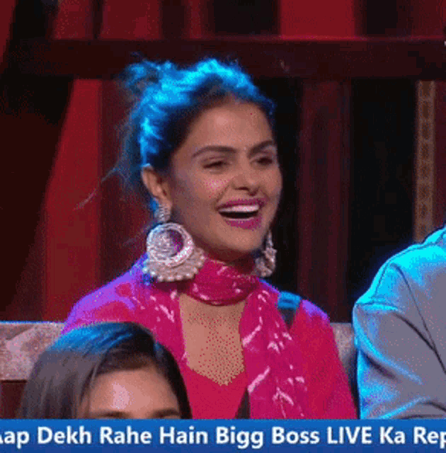 a woman in a pink dress is laughing in front of a banner that says " dekh rahe hain bigg boss live ka rep "