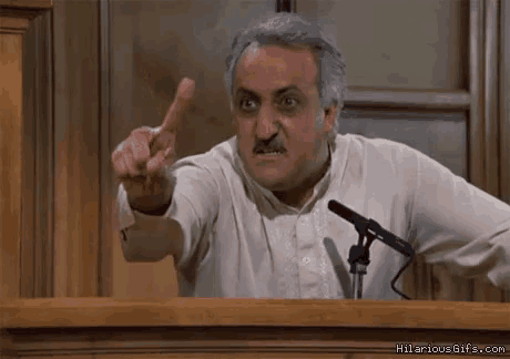 a man is giving a speech in front of a microphone and pointing his finger at the camera .