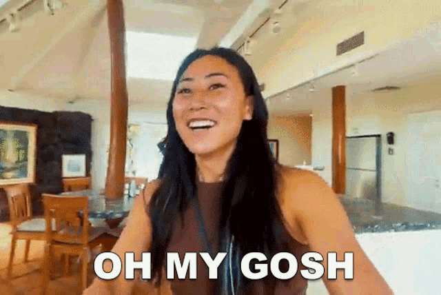 a woman is standing in a living room and smiling with the words oh my gosh on her face .