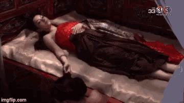 a woman in a red dress is laying on a bed with a man standing next to her .
