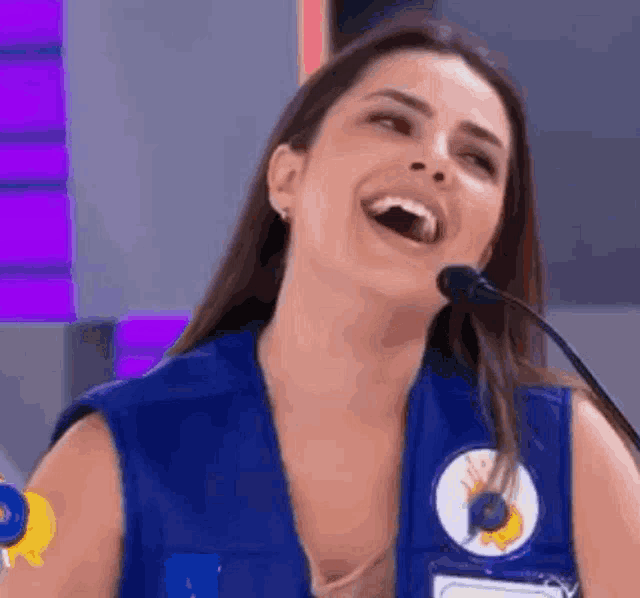 a woman in a blue vest is laughing into a microphone while wearing a name tag .