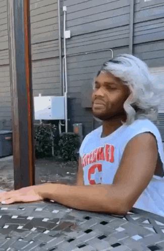 a man with a wig is sitting at a table with his arms outstretched .