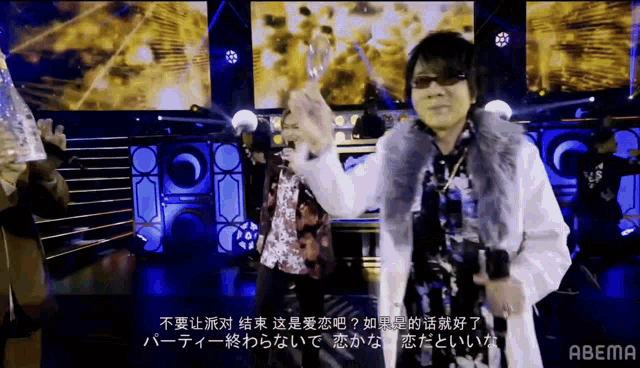 a man wearing sunglasses and a fur coat sings into a microphone on a stage with abema in the corner