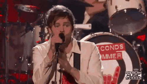 a man is singing into a microphone with a drum set in the background .