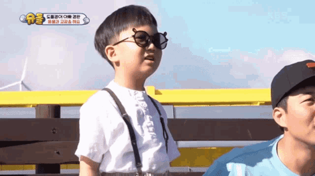 a little boy wearing suspenders and sunglasses stands next to a man wearing a baseball cap ..
