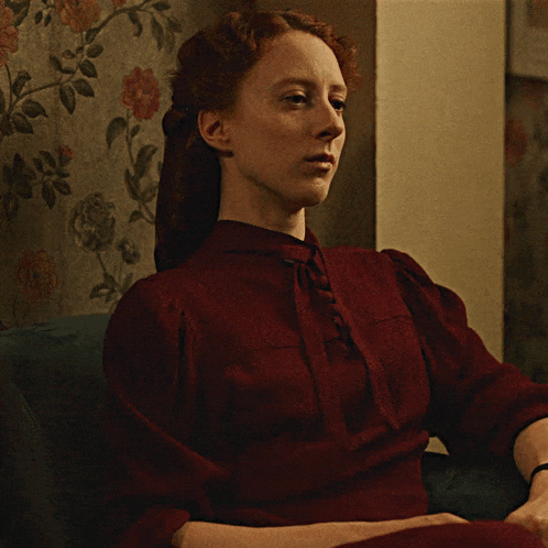 a woman in a red dress sits in front of a floral wallpaper