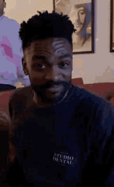 a man with a beard is sitting on a couch in a living room wearing a black shirt .