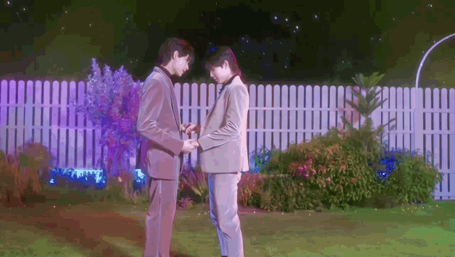 two men in suits stand next to each other in front of a white picket fence