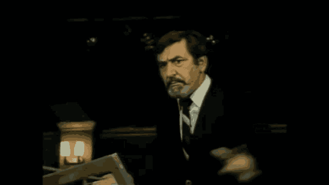 a man in a suit and tie is standing in a dark room holding a book and talking .
