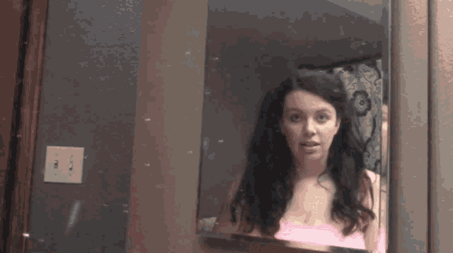 a woman in a pink tank top is looking at herself in a bathroom mirror