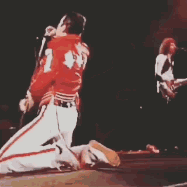 a man in a red jacket is singing into a microphone on a stage while another man plays a guitar .