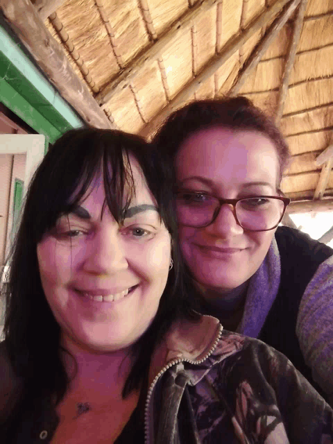 two women are posing for a picture with a thatched roof behind them
