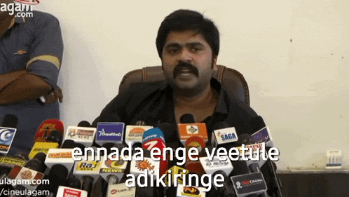 a man stands in front of a bunch of microphones with the words " ennada enge veetule adikiringe " above him