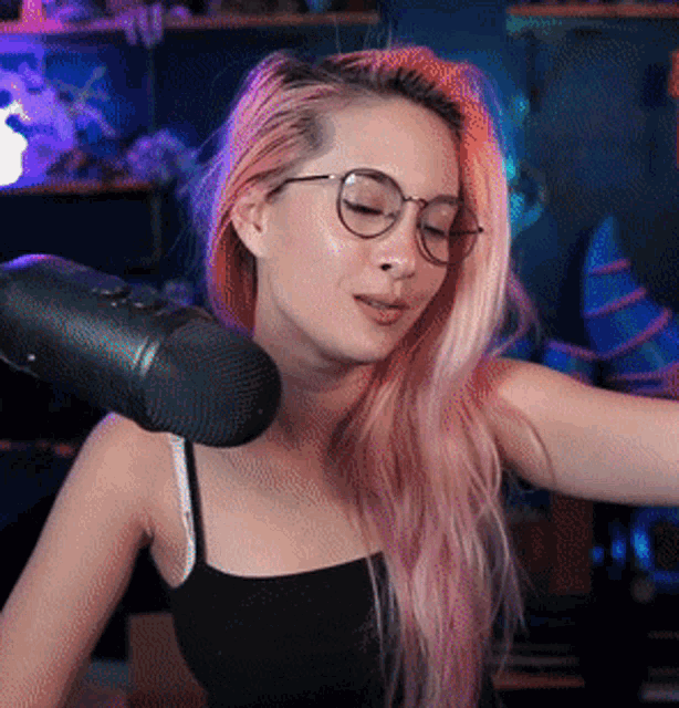 a woman with pink hair and glasses stands in front of a microphone with her eyes closed