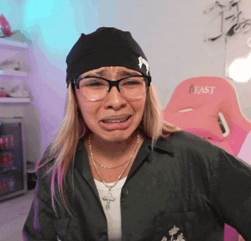 a woman wearing glasses and a black hat is crying in front of a pink chair that says beast on it