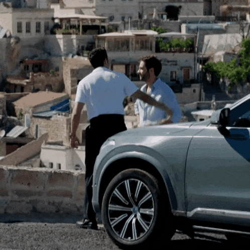 two men are standing next to a silver car in front of a city