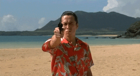 a man in a hawaiian shirt is pointing a gun at the camera on the beach .